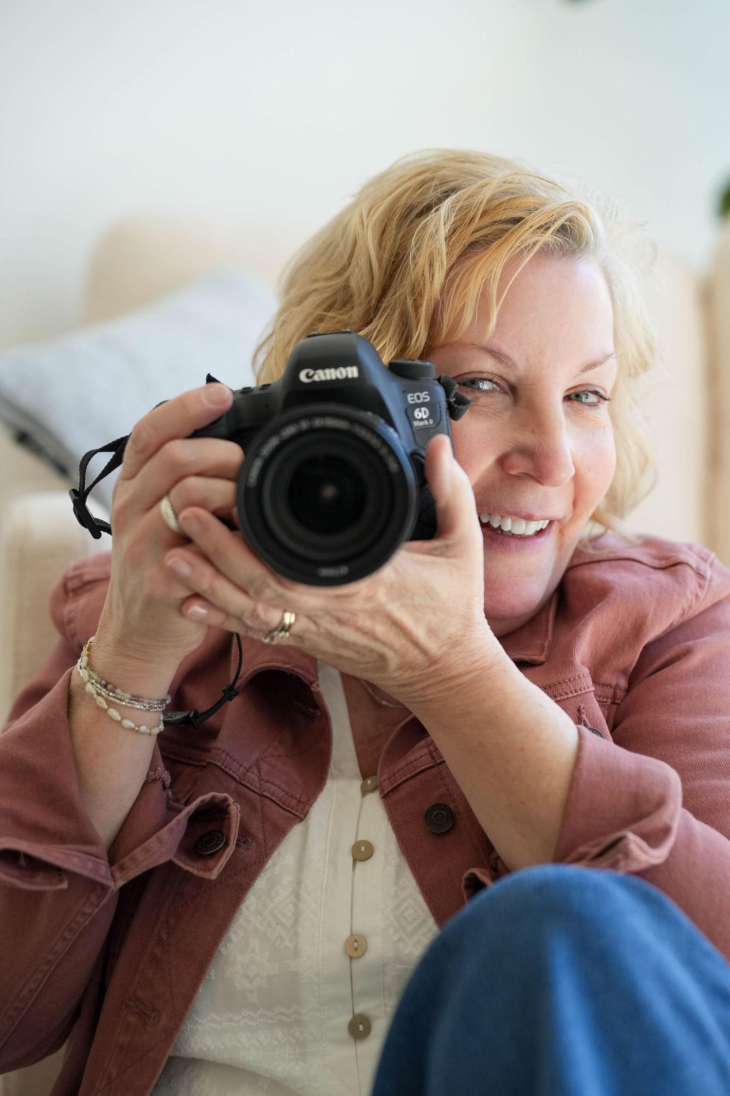 Smiling blonde woman holding a Canon EOS DSLR camera up to take a photo while sitting on a couch indoors.