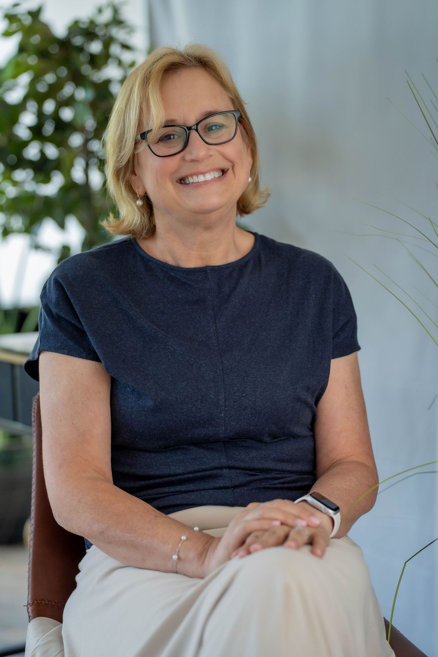 Smiling middle-aged woman with blonde hair and glasses, wearing a navy t-shirt, seated indoors near green plants.