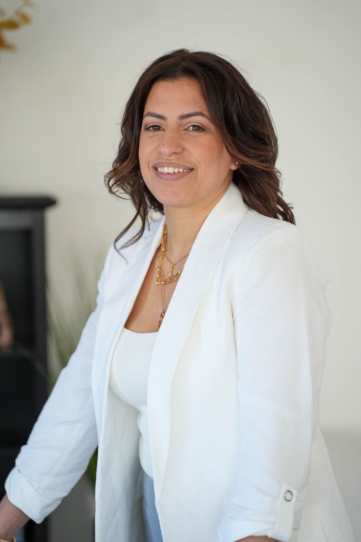 Smiling woman in white blazer and gold necklace posing indoors with a bright, modern background.