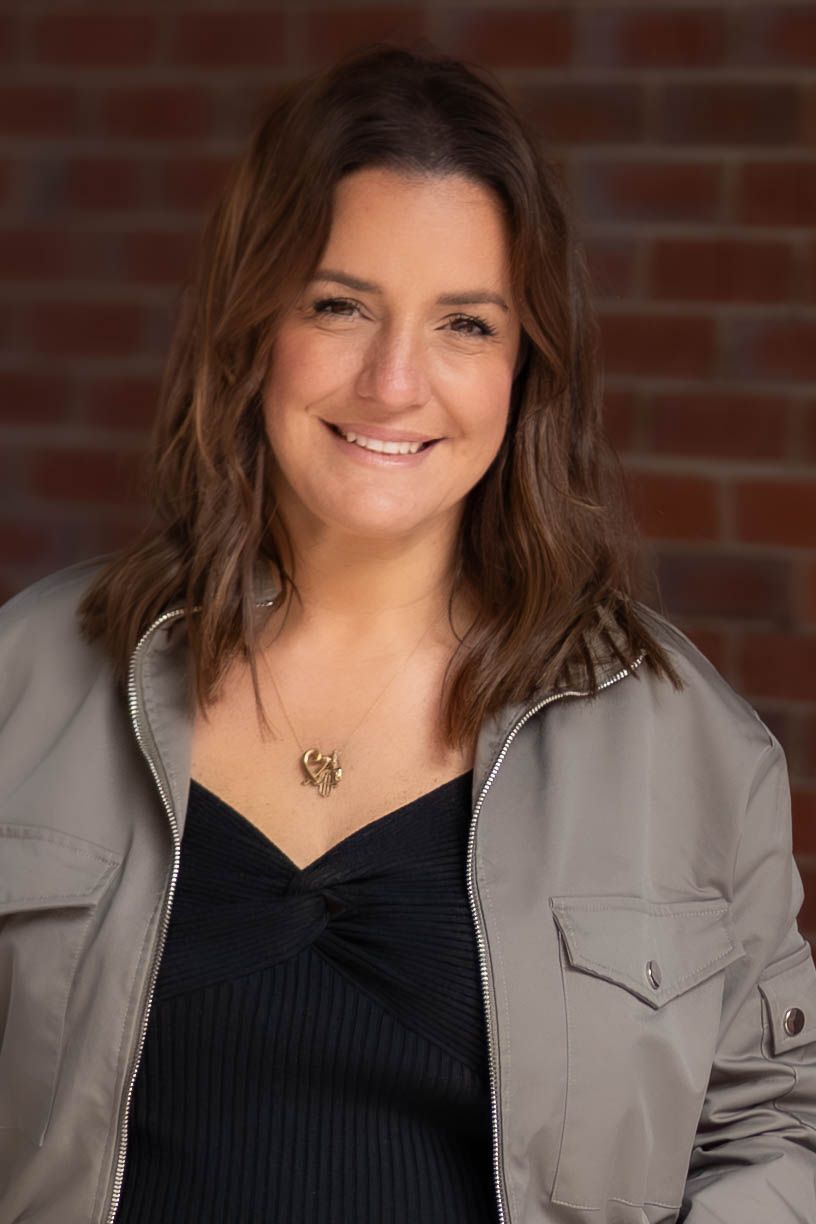 Smiling woman with brown hair wearing a gray jacket and black top, posing in front of a brick wall.