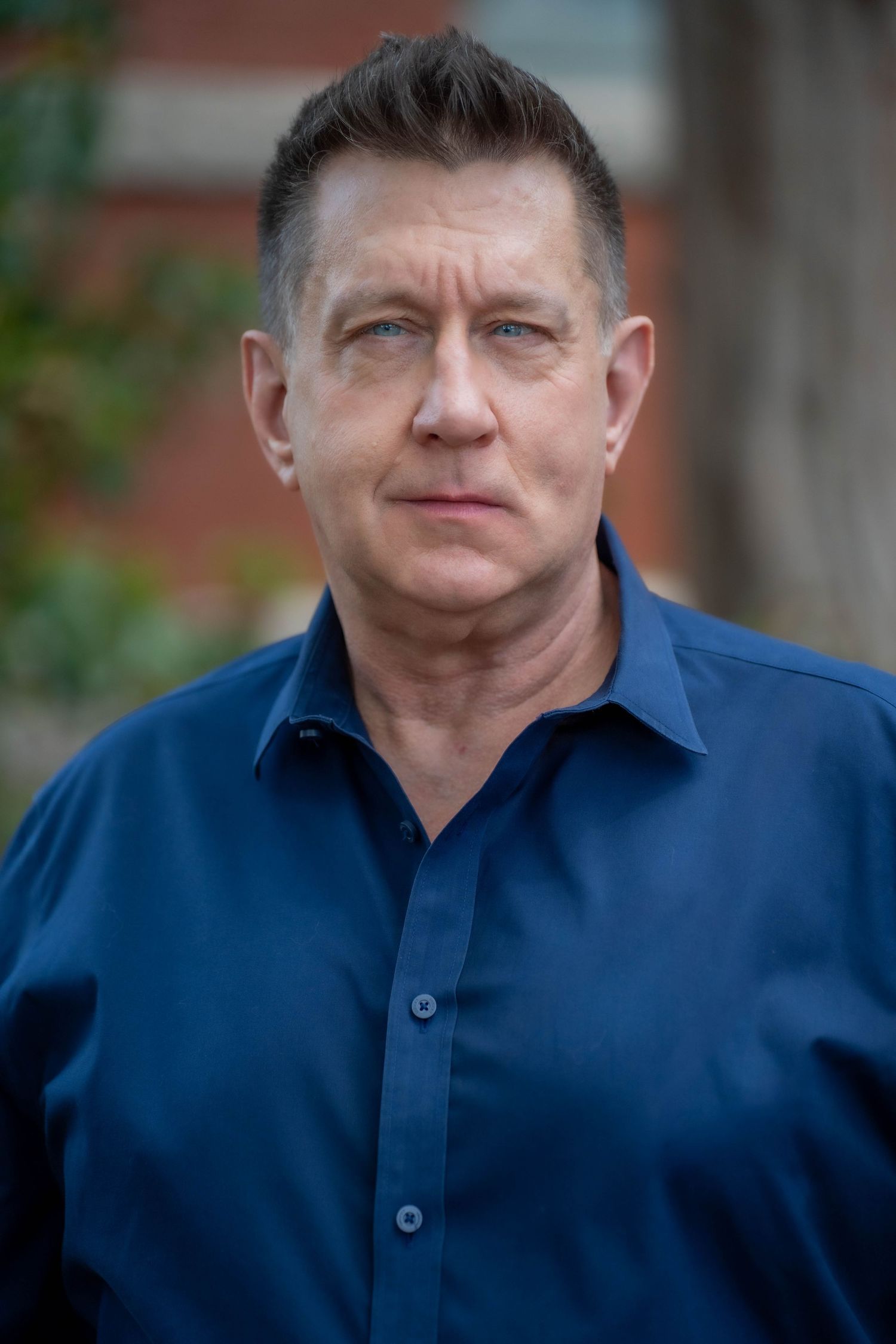 Middle-aged man in navy blue button-up shirt poses outdoors with serious expression, brick building in background.