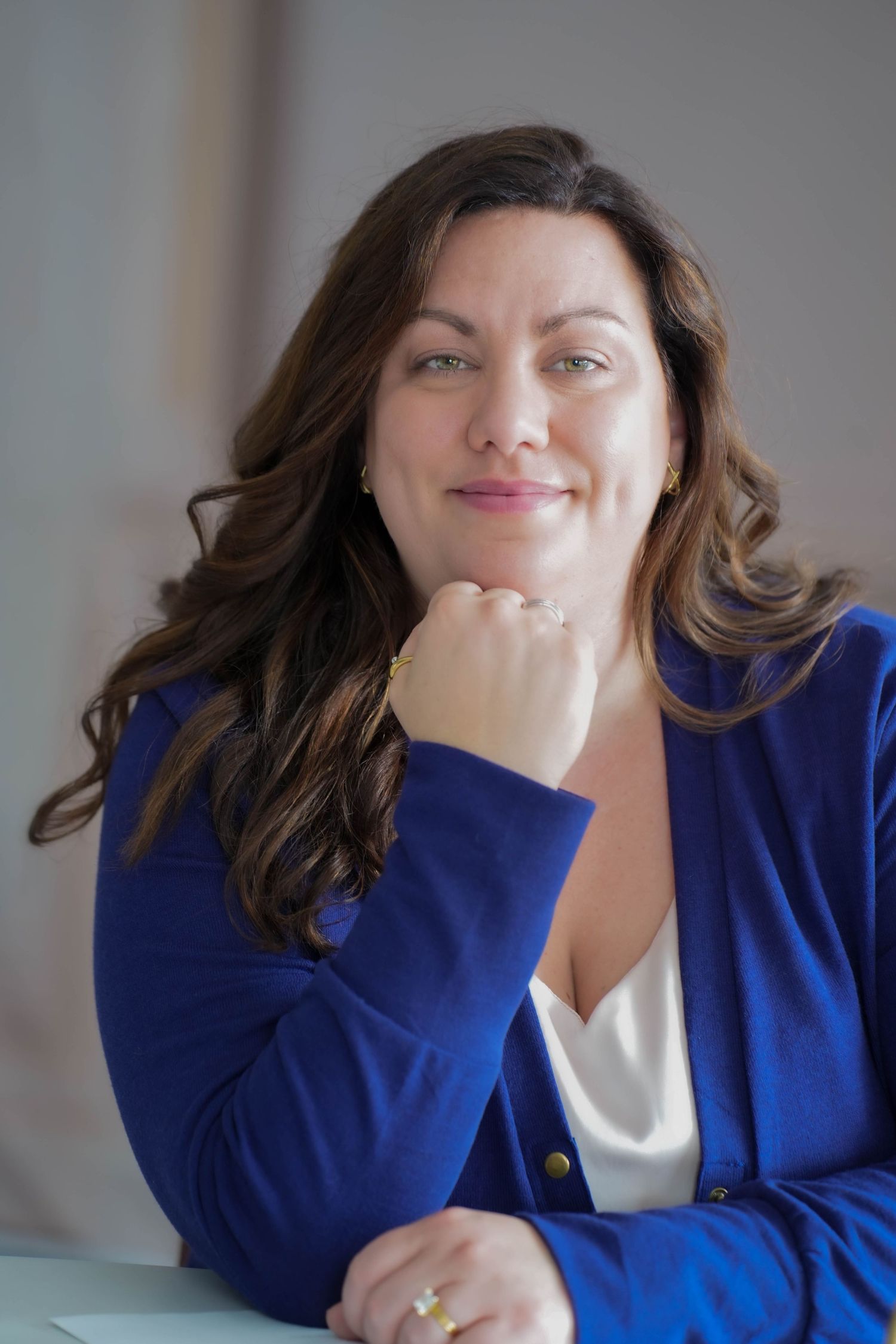 Professional woman in blue blazer smiling confidently, resting chin on hand in a modern office setting.