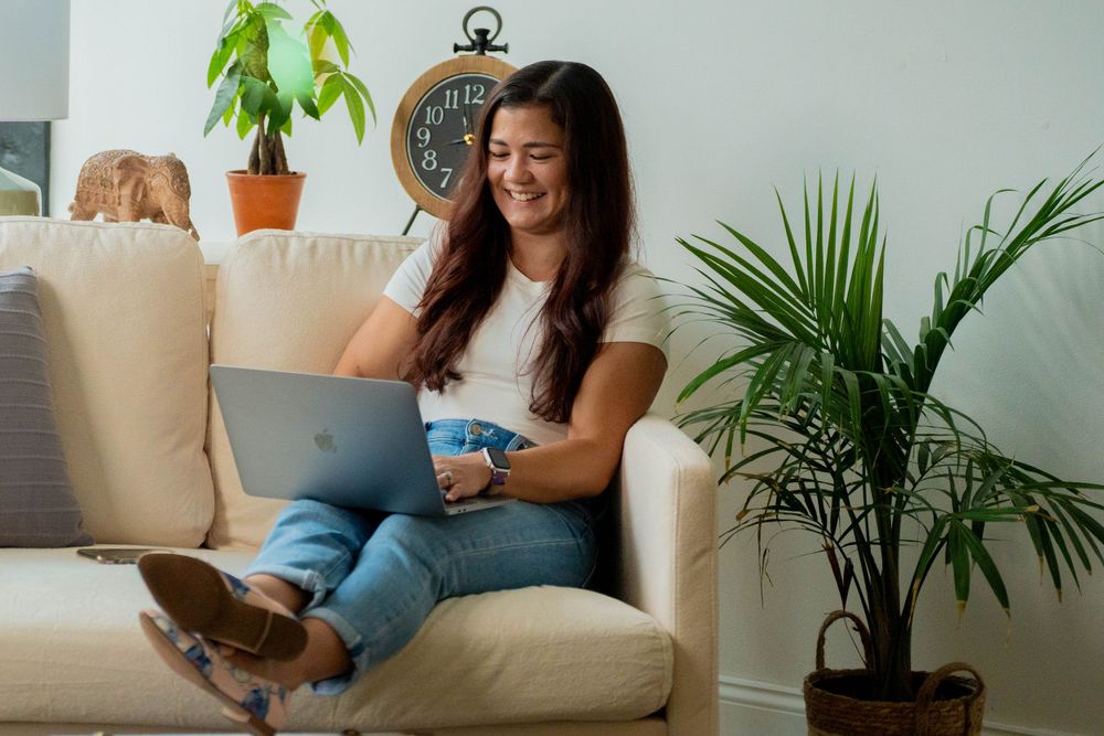 Woman relaxing on beige couch with laptop in living room with plants.
