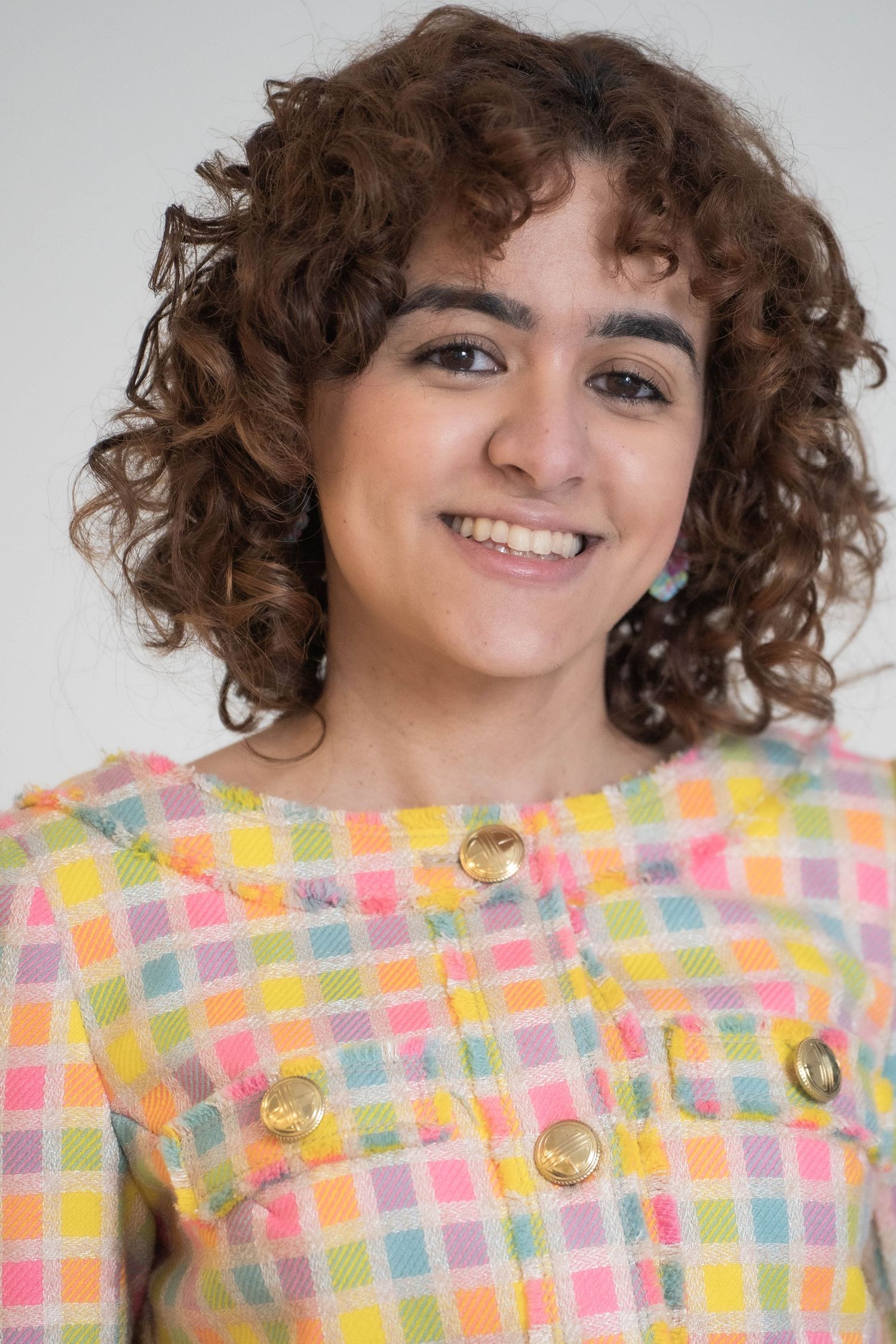 Smiling young woman with curly brown hair wearing a colorful pastel plaid jacket with gold buttons.