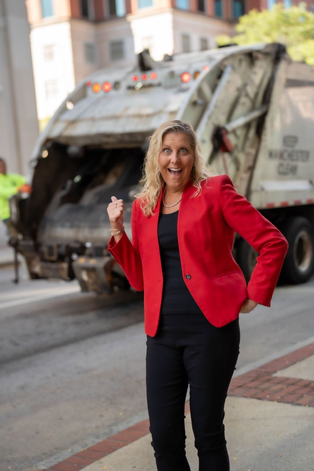 Woman in red blazer and black outfit posing confidently near garbage truck on city street.