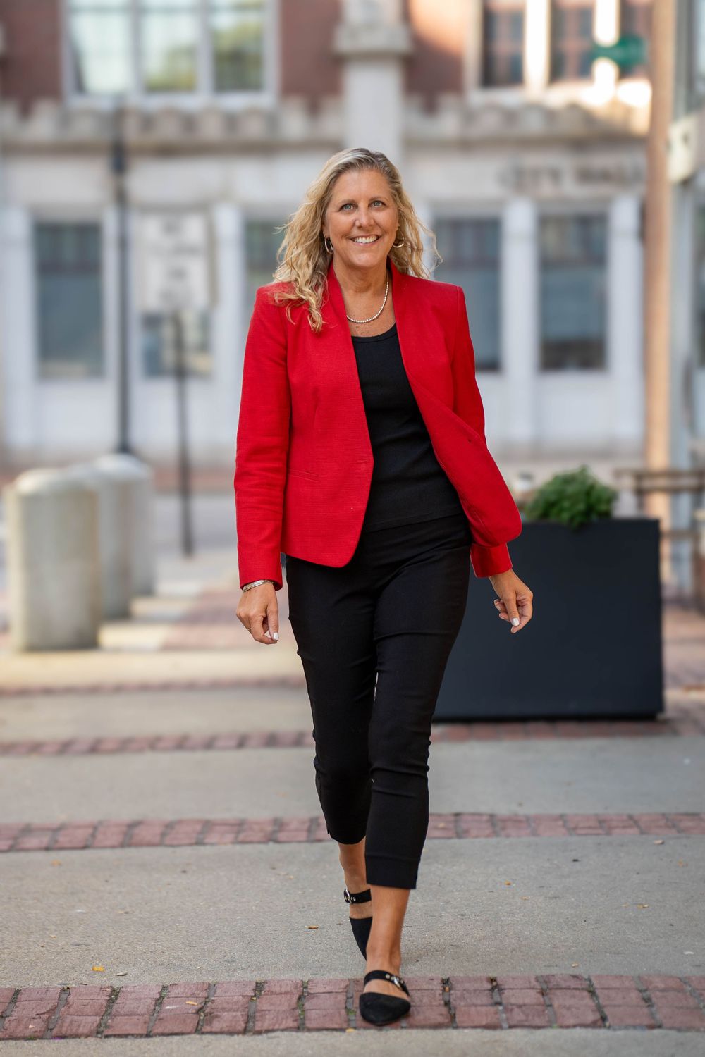 Woman in bright red blazer and black pants walking on brick sidewalk downtown.