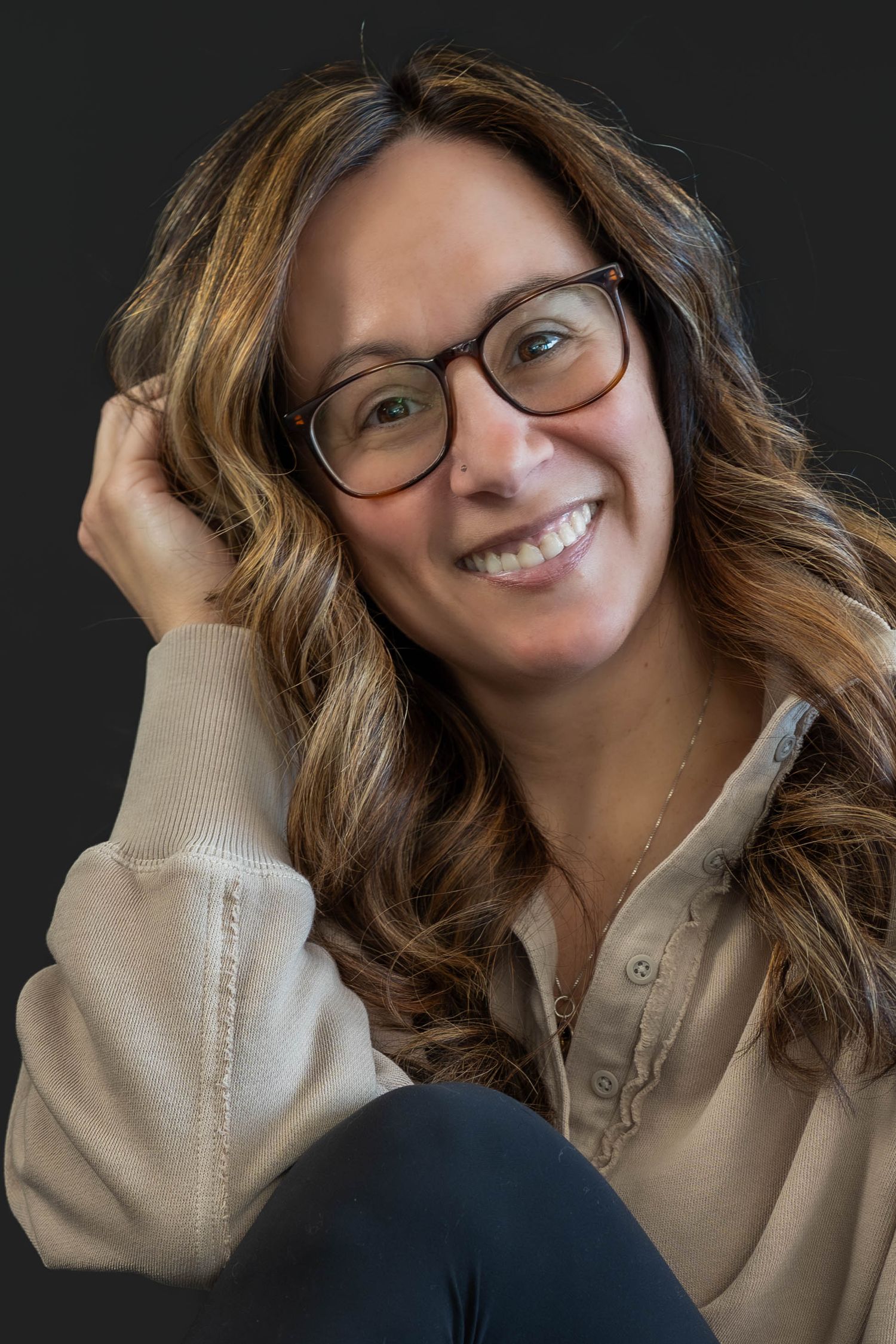 Smiling woman with glasses and wavy hair posing casually against dark background.