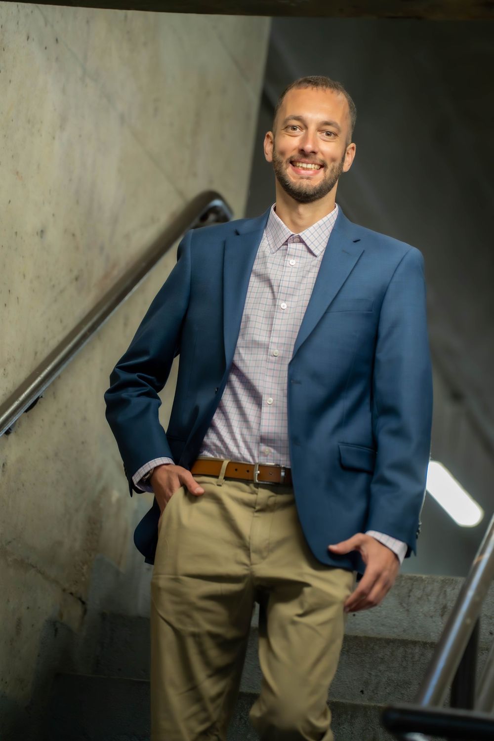 Professional poses in navy blazer and khaki pants against concrete background.