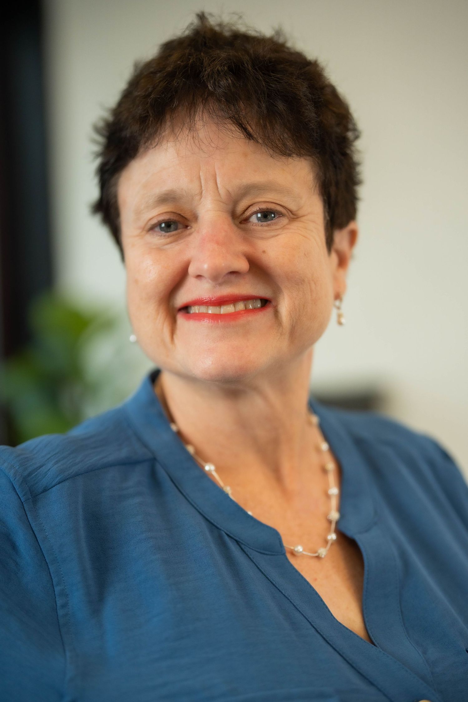 Smiling middle-aged woman with short dark hair wearing a blue blouse and pearl necklace in a professional headshot.