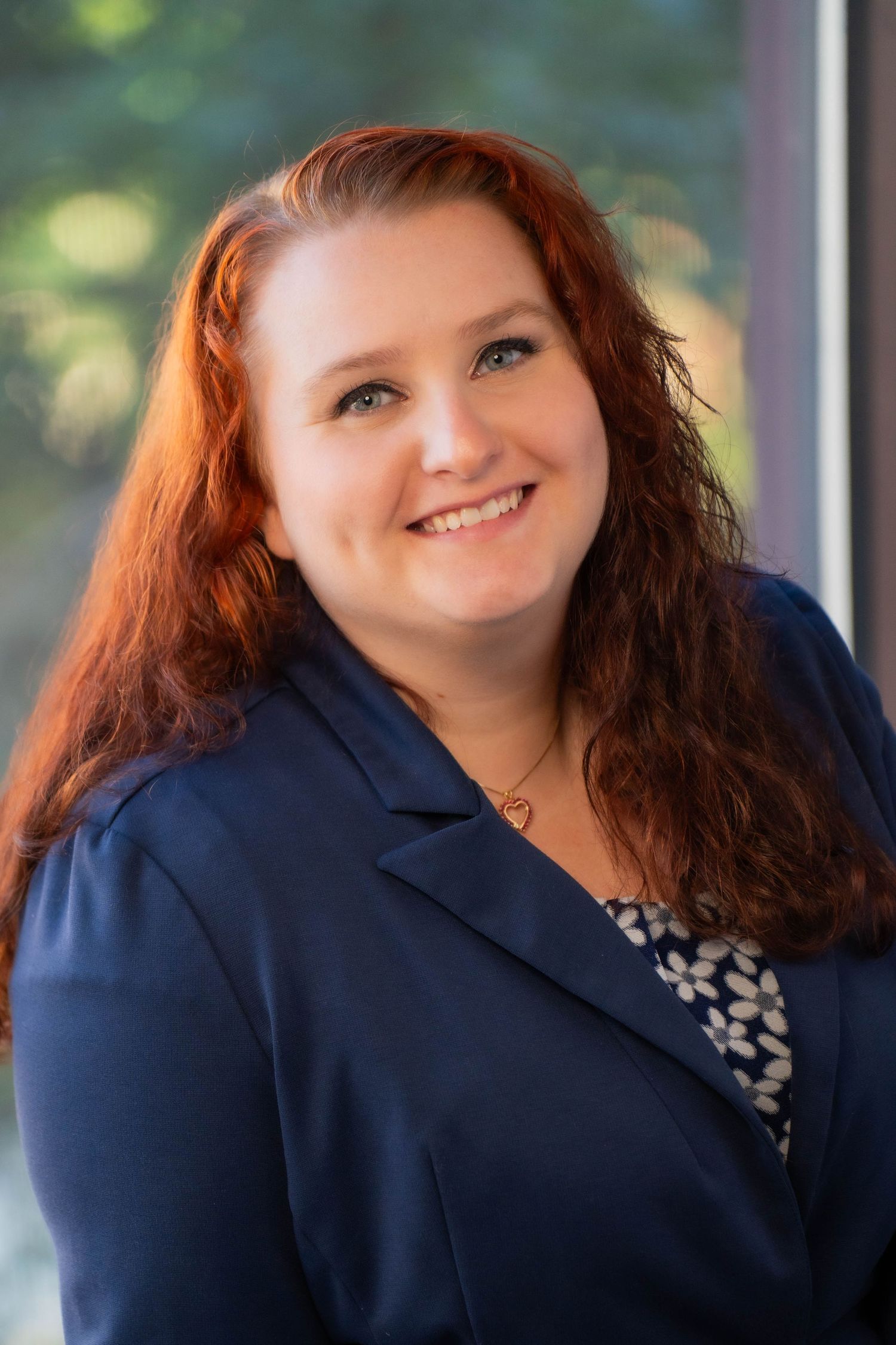 Professional headshot of a smiling woman with red hair wearing a navy blazer, photographed near a window.