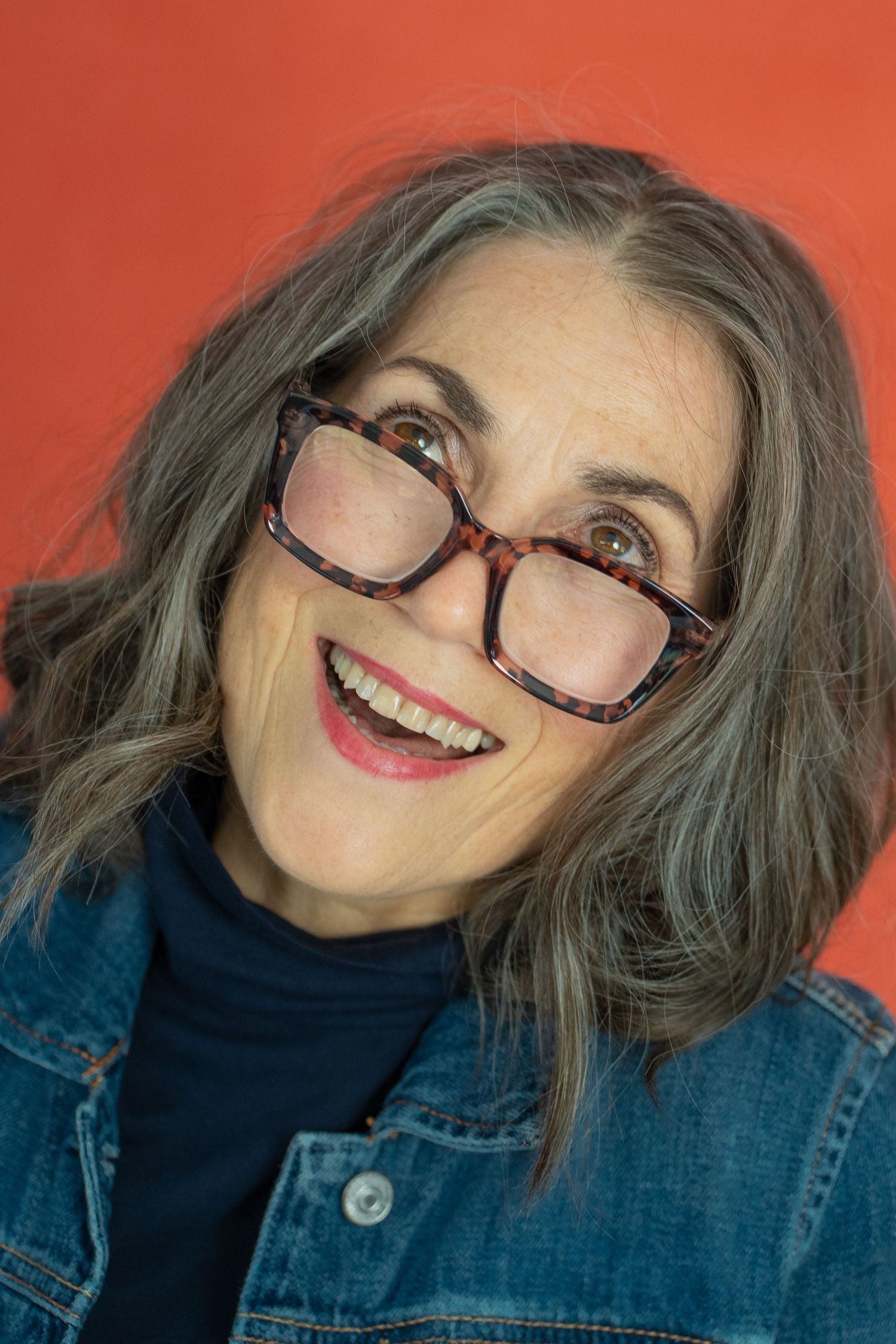 Smiling middle-aged woman with gray hair and tortoiseshell glasses wearing a denim jacket against a red background.