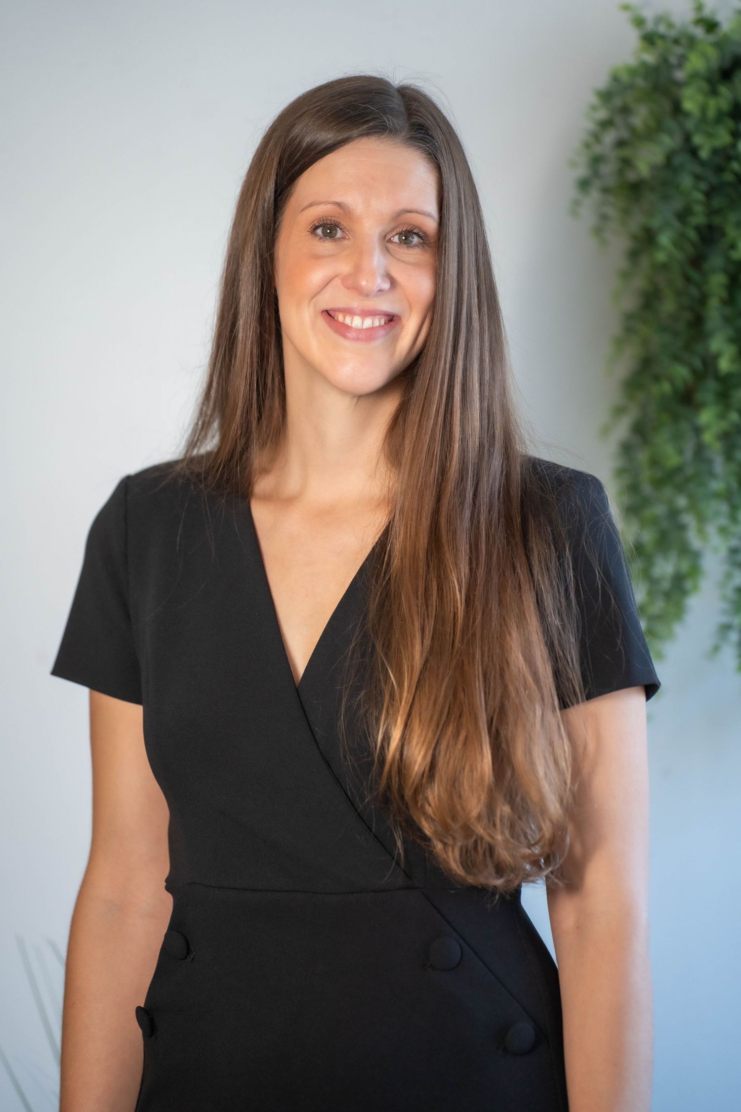 Woman in black wrap dress smiling, long brown hair, professional headshot with green plant background.