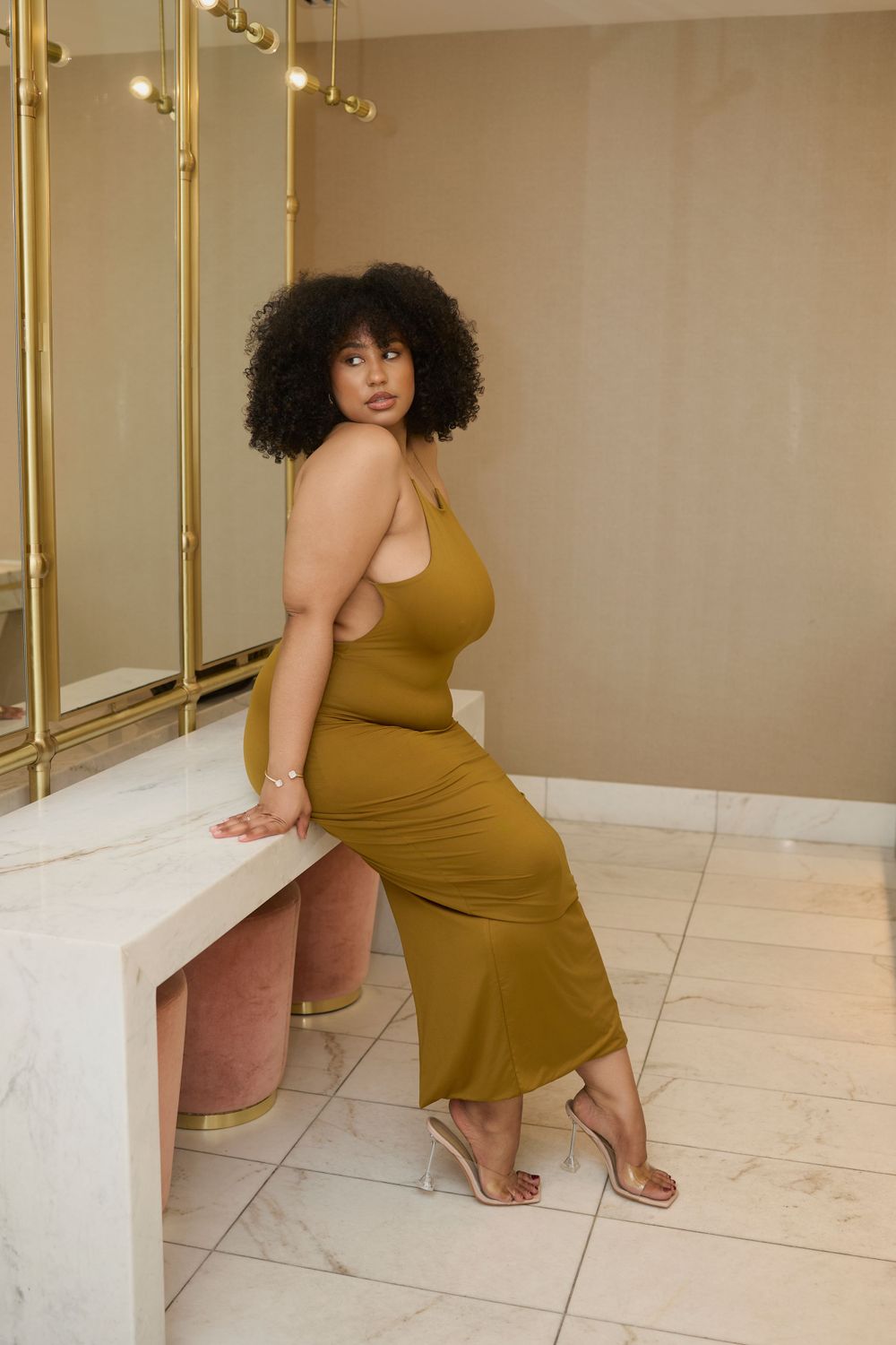 Model in mustard-colored dress poses against beige interior with gold accents.