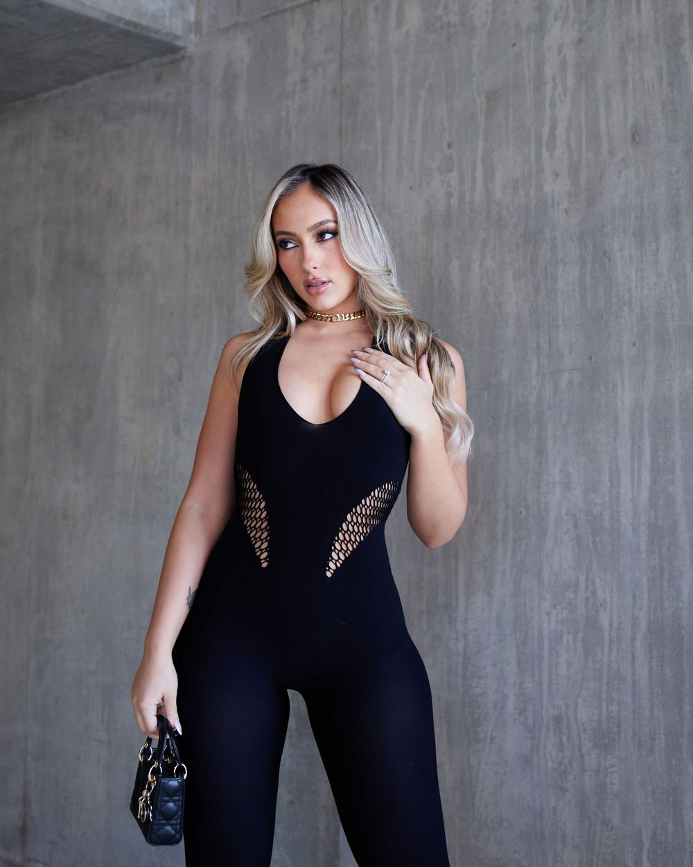 A black fitted jumpsuit with mesh cutouts shown against an industrial concrete background.