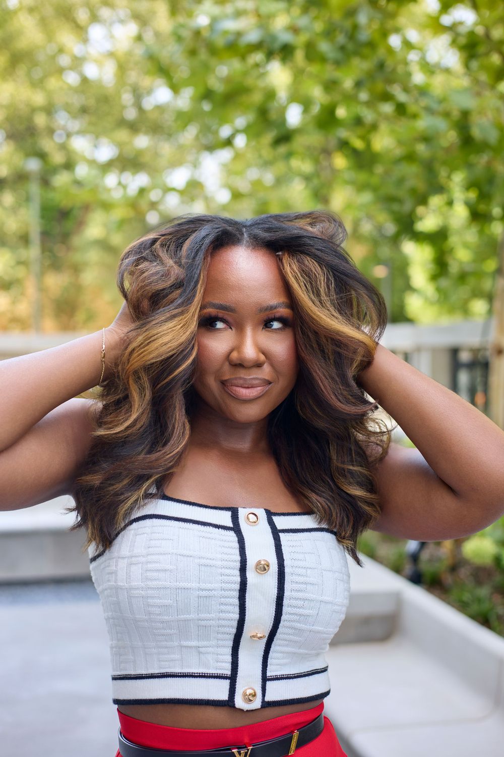 A carefree pose in a white cropped top with black trim against a leafy outdoor background.