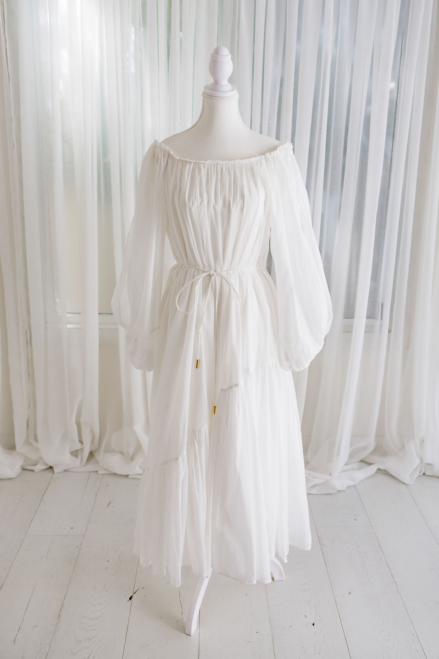 Full-length white cotton dress with billowing sleeves on mannequin against sheer curtains.
