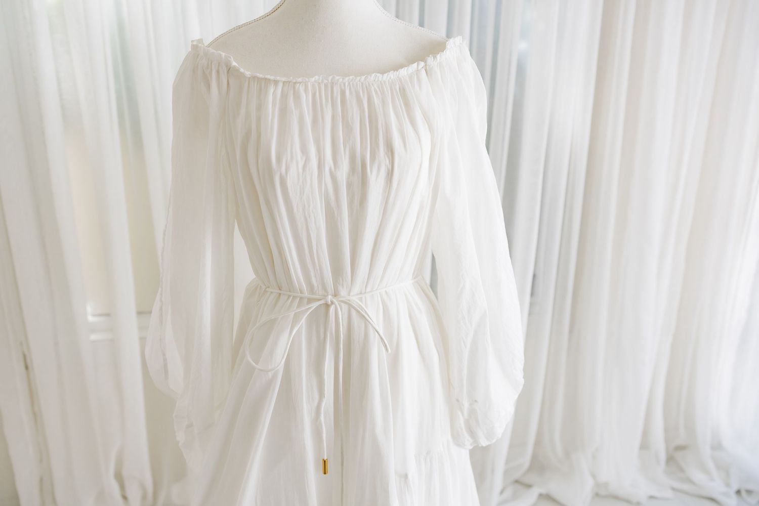 Ethereal white cotton dress with gathered neckline and billowing long sleeves on display against white curtains.