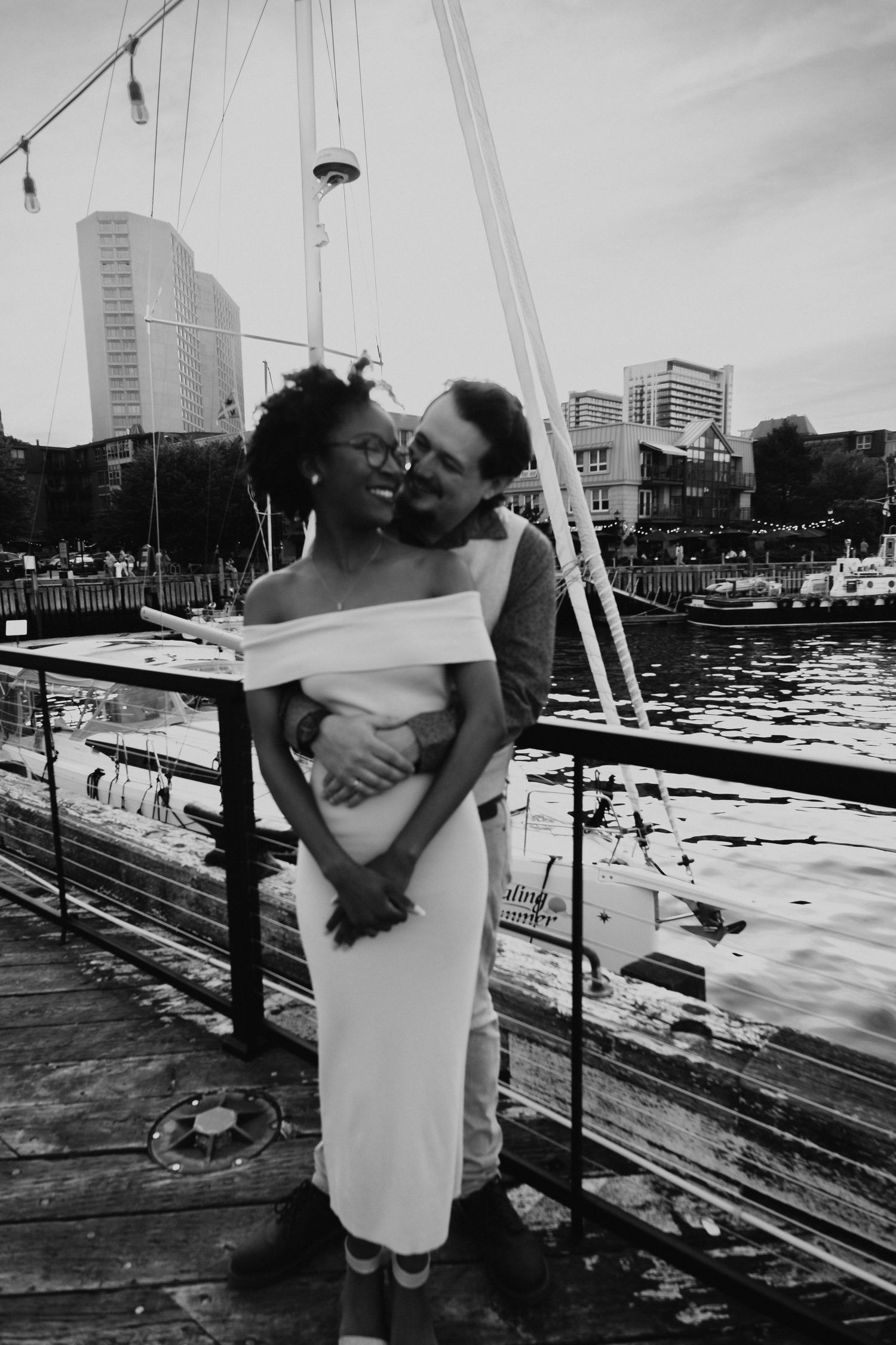 Romantic couple embracing on a waterfront dock with city skyline in background.