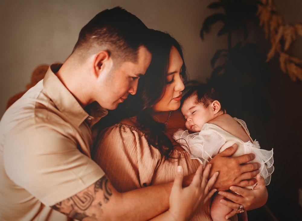A warm-toned intimate family moment with parents embracing their sleeping newborn baby.