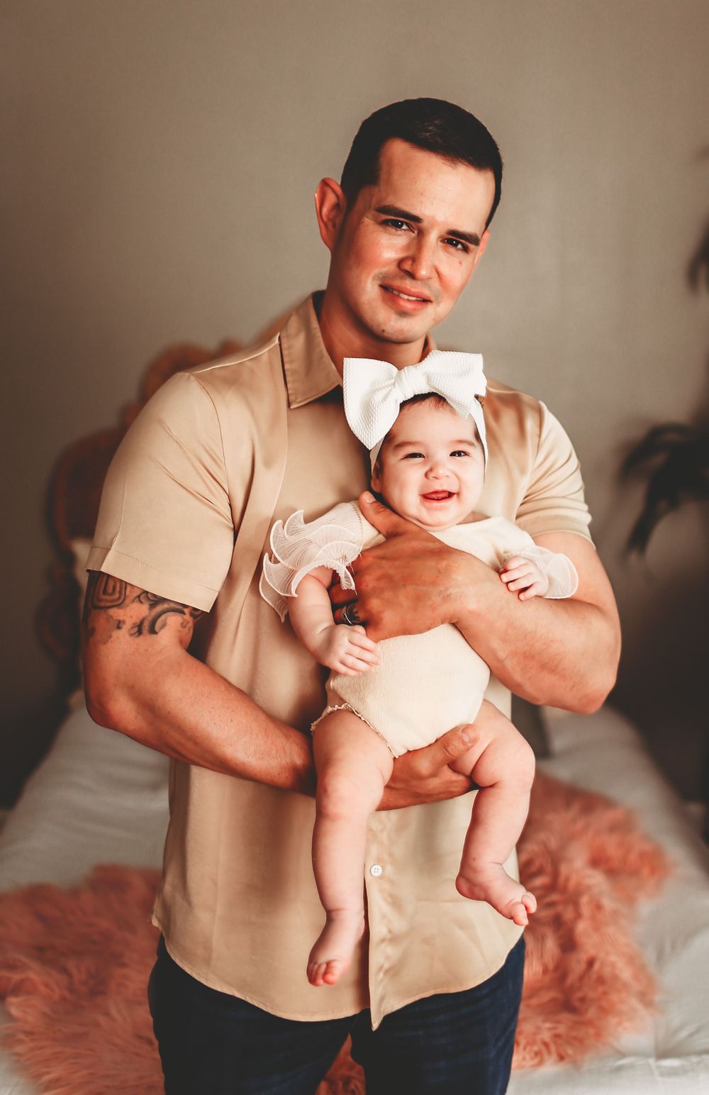 Smiling dad in beige shirt holding happy baby in white outfit sitting on pink furry blanket.