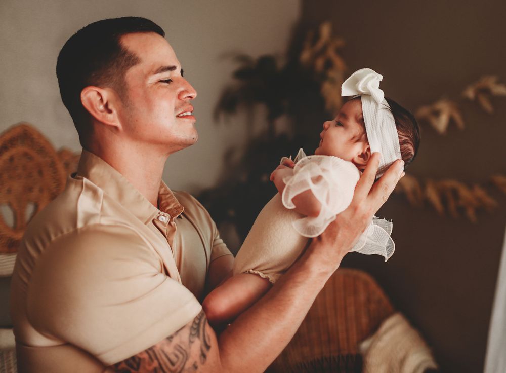 daddy in beige clothing holds a small baby dressed in white during an intimate indoor moment.