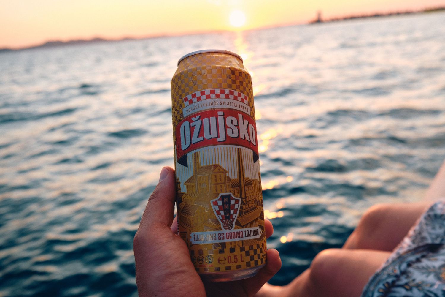 Hand holding Ozujsko beer can against sunset ocean backdrop.
