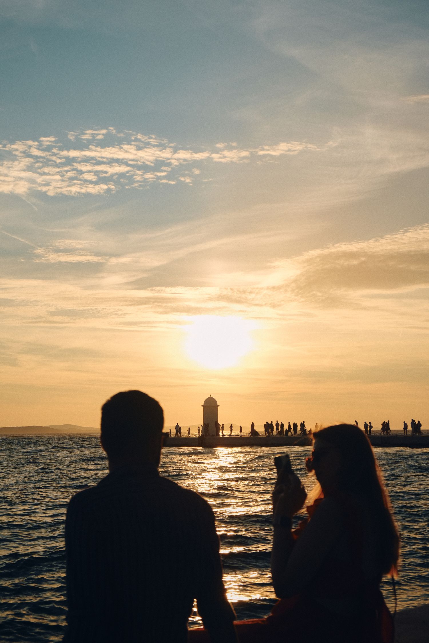 Silhouettes of people watching sunset by a lighthouse with crowd gathered along the shore.