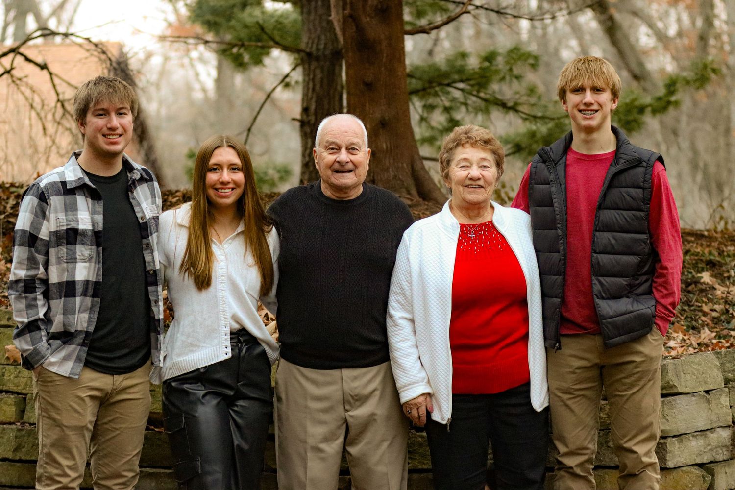 Family group of five poses together in casual attire against stone wall and trees.