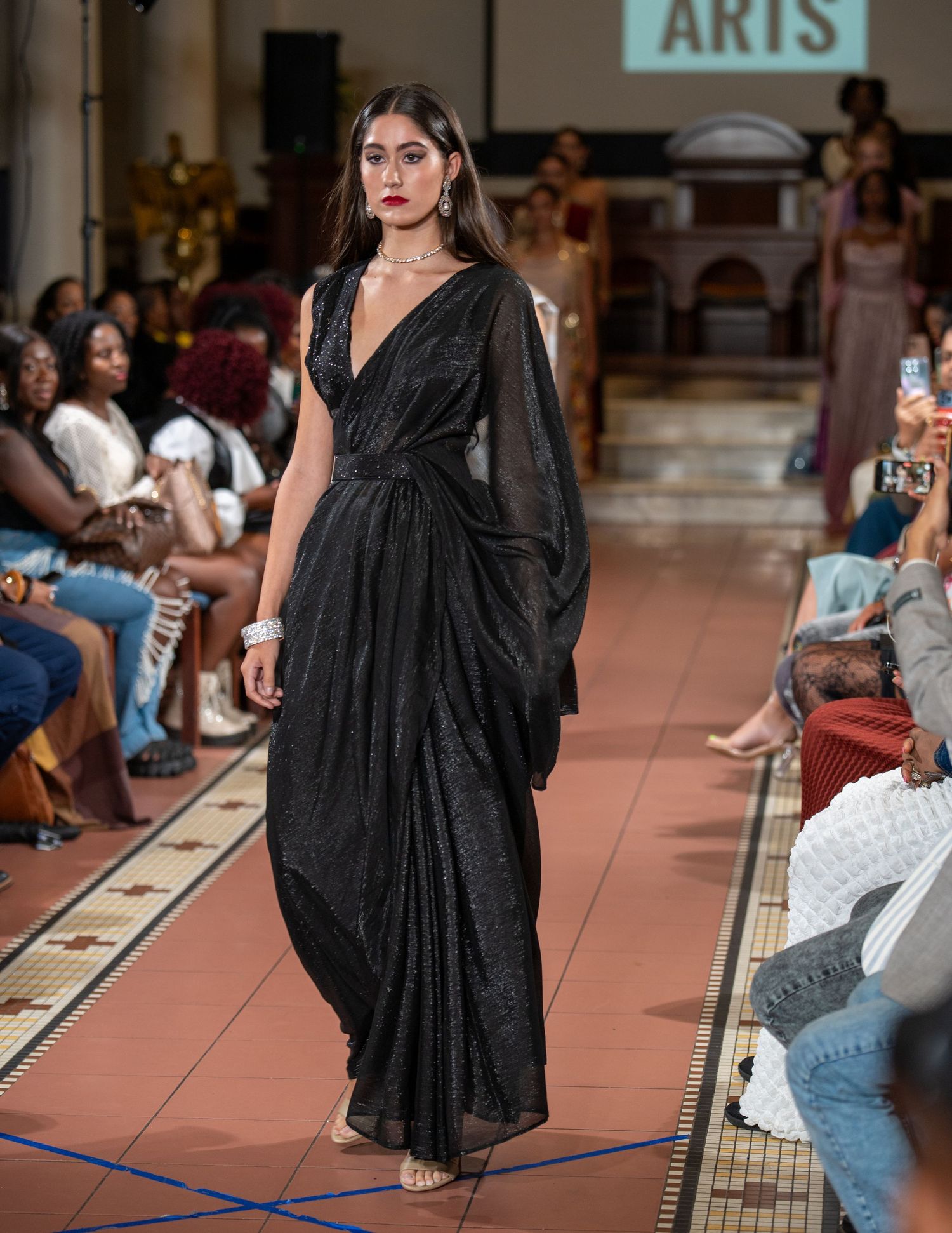 Dramatic black draped evening gown showcased on fashion runway with audience.