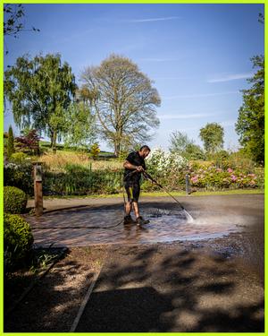 A person in dark clothing pressure washes a driveway on a sunny day surrounded by lush greenery and trees.