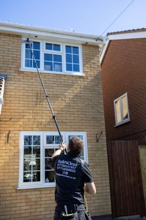 Professional window cleaner uses a water-fed pole system to clean the upper windows of a yellow brick house.