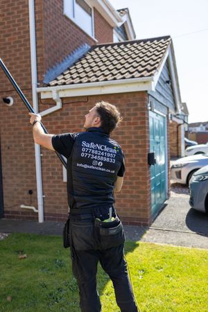 A window cleaner in a SafeInClean company uniform uses an extension pole to clean windows on a brick house.