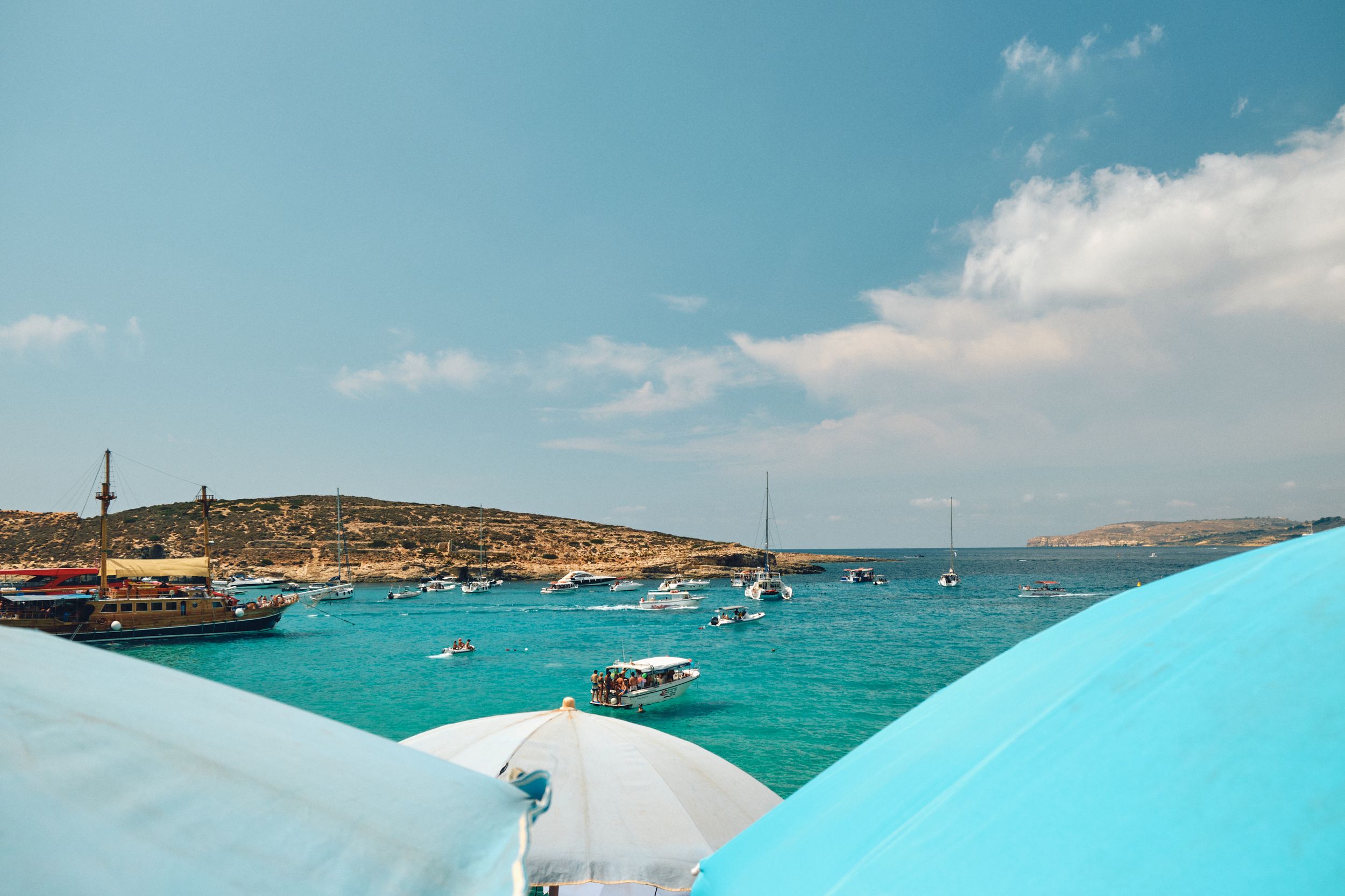 Aerial view of boats anchored in crystal clear turquoise waters of Blue Lagoon in Malta with beach umbrellas visible.