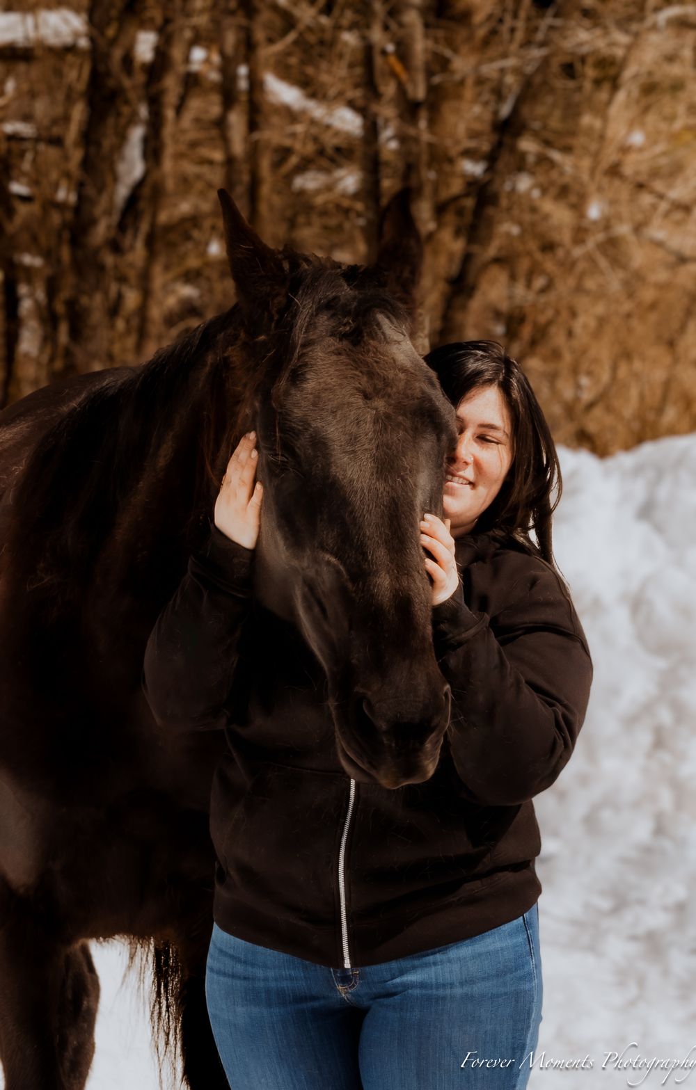 A tender moment between a dark horse and person in a winter forest setting with snow on the ground.