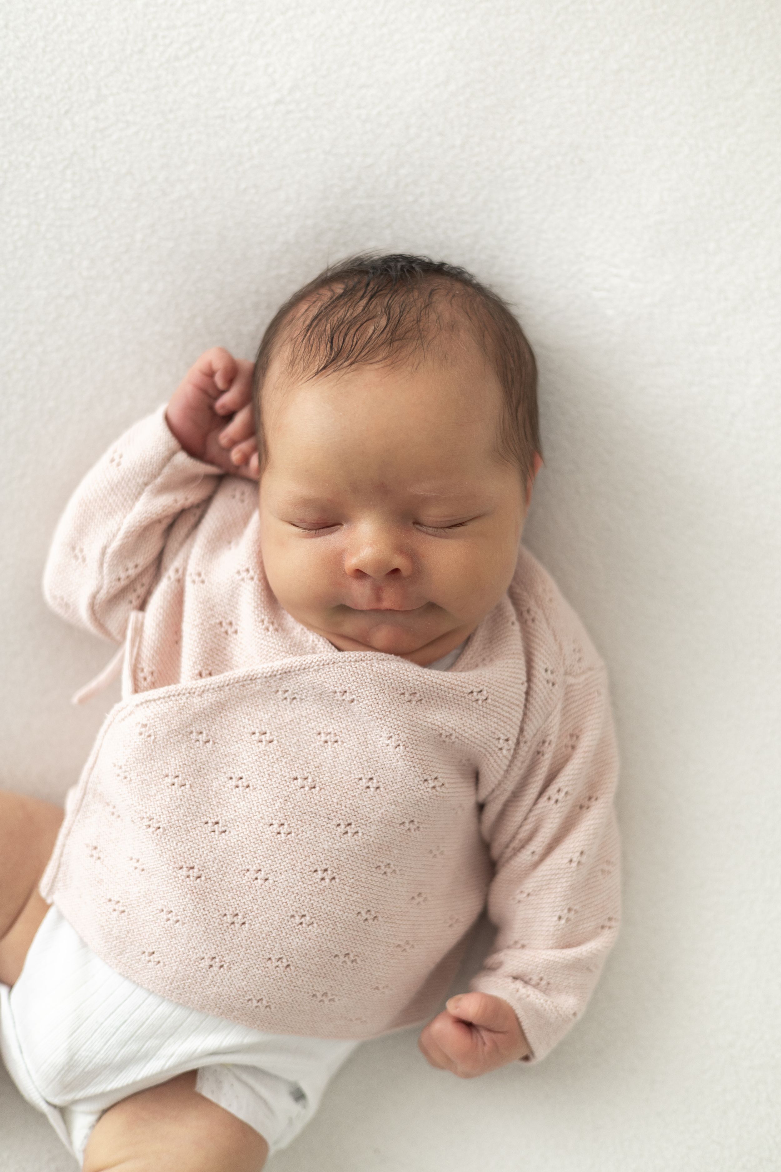 Sleeping newborn baby in pink knit jumper lying on white blanket with hand raised near face nature newborn photographer Nottingham