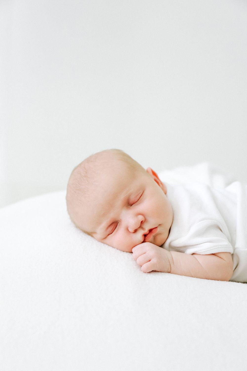 A peaceful baby sleeps soundly on white bedding in a peaceful and serene pose.