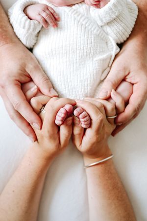 Adult hands gently cradle tiny baby feet against soft white knit sweater background.