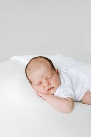 A sleeping newborn baby dressed in white lies peacefully on white bedding.