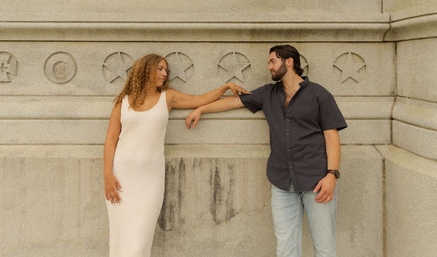 Playful couple posing against ornate stone wall in casual attire.