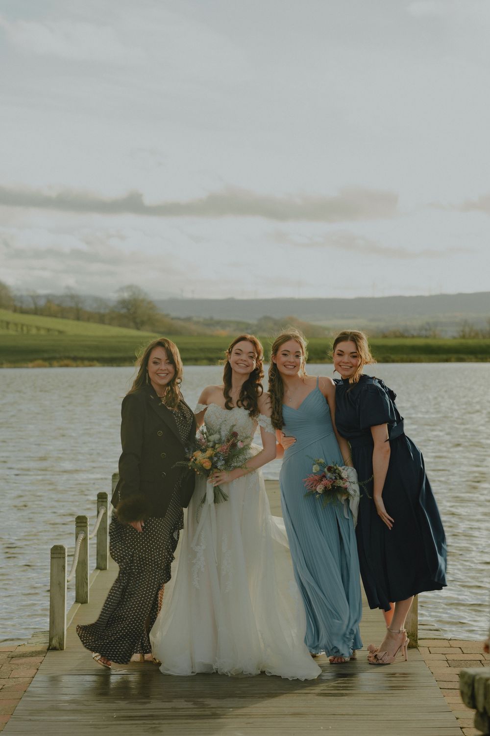 Bride with bridesmaids in blue dresses posing on a lakeside dock at golden hour.