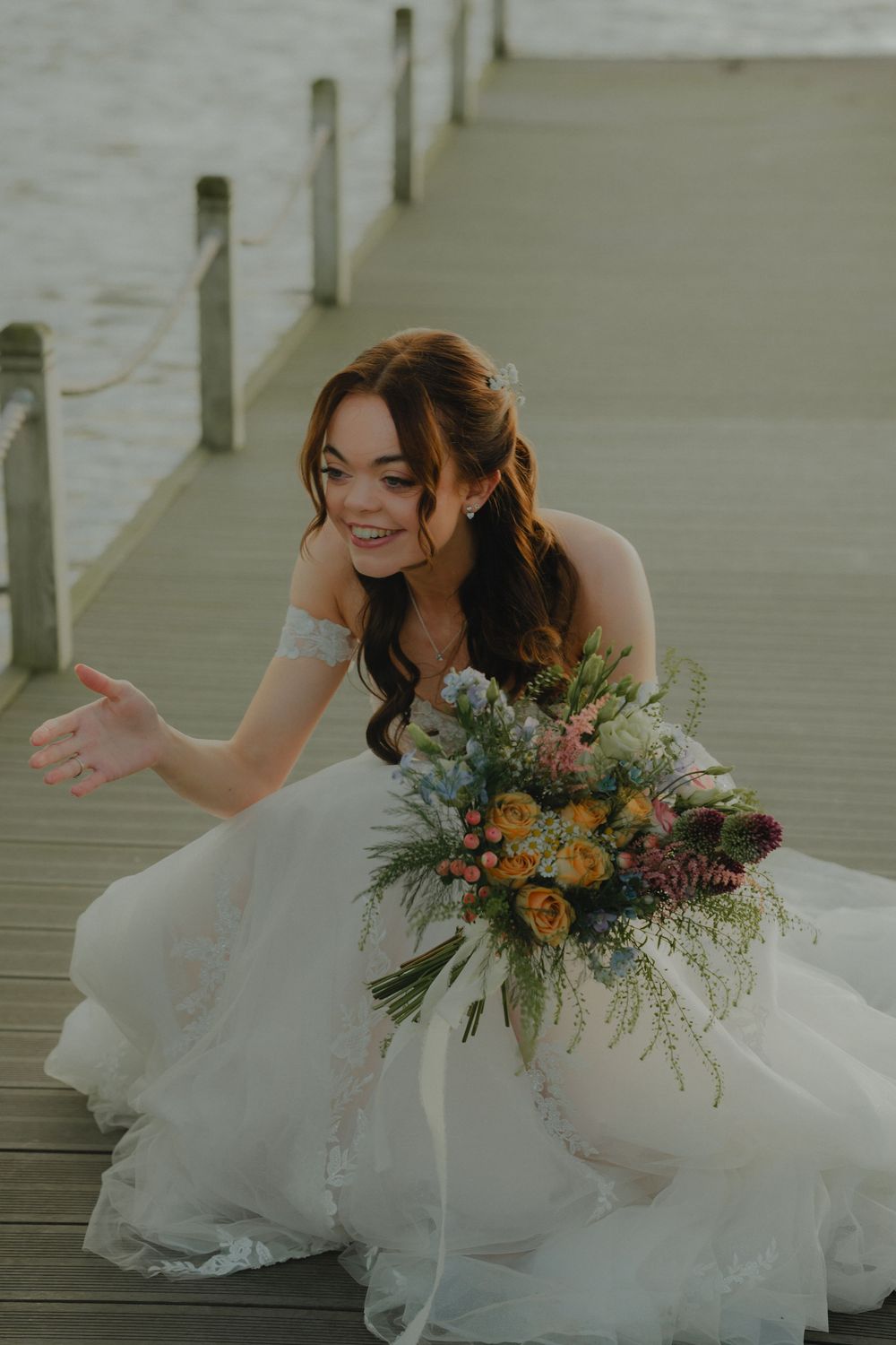 Smiling bride in white lace gown sitting on dock holding colorful wildflower bouquet.