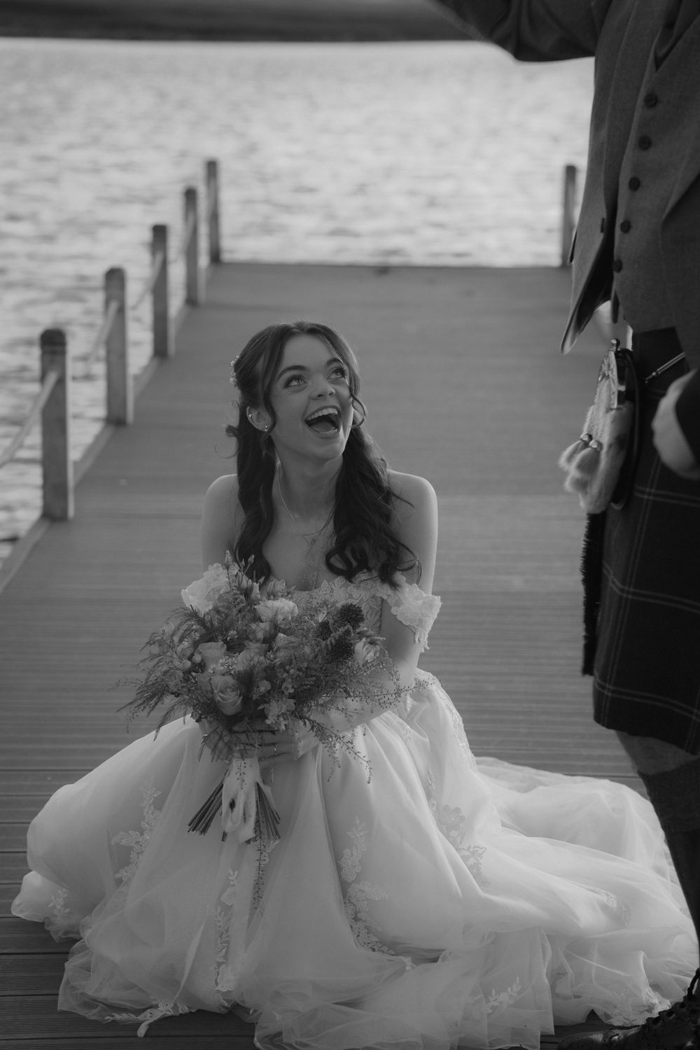 Joyful bride in a white ball gown holding a bouquet, laughing on a wooden dock by the water.