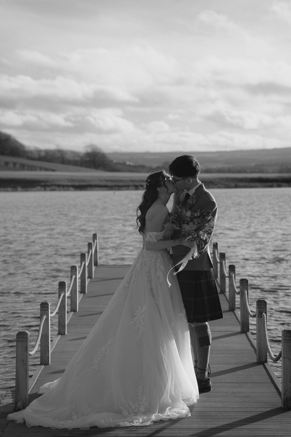 Black and white photo of bride and groom in kilt sharing romantic moment on lake dock.