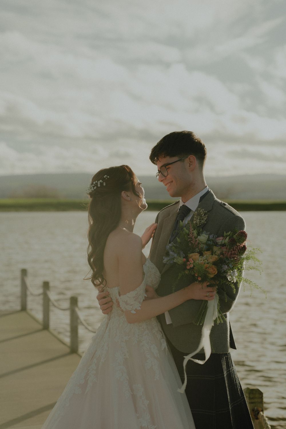 Bride and groom share a romantic moment on a lakeside dock, bride holding a wildflower bouquet in wedding attire.