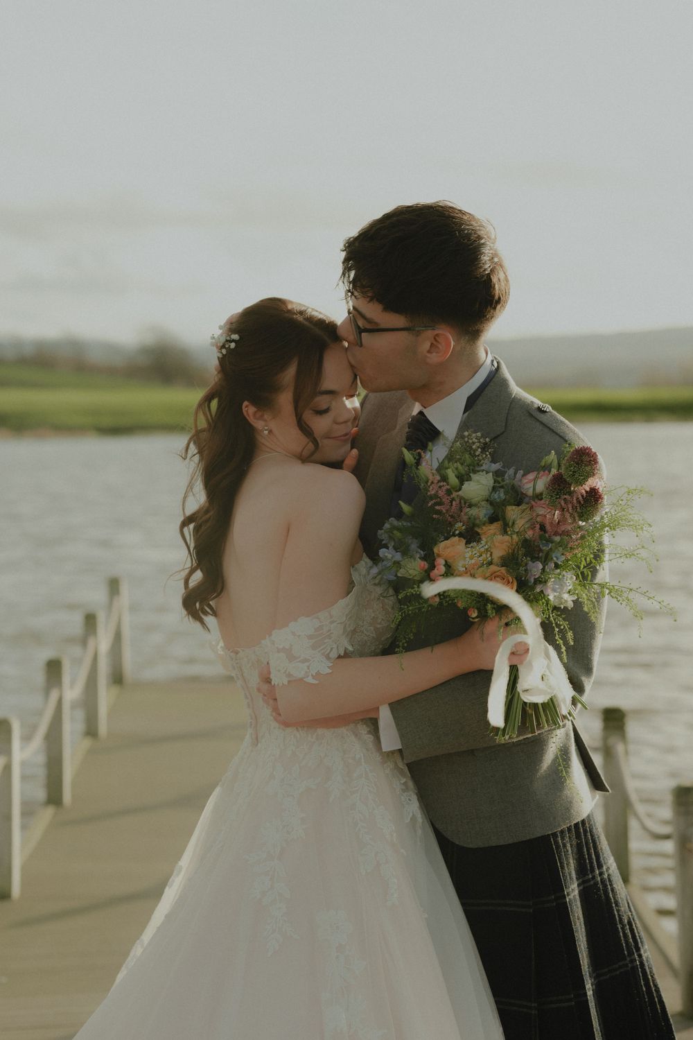 Groom in tartan kilt kissing bride on cheek on lakeside dock, bride holding colorful wedding bouquet.