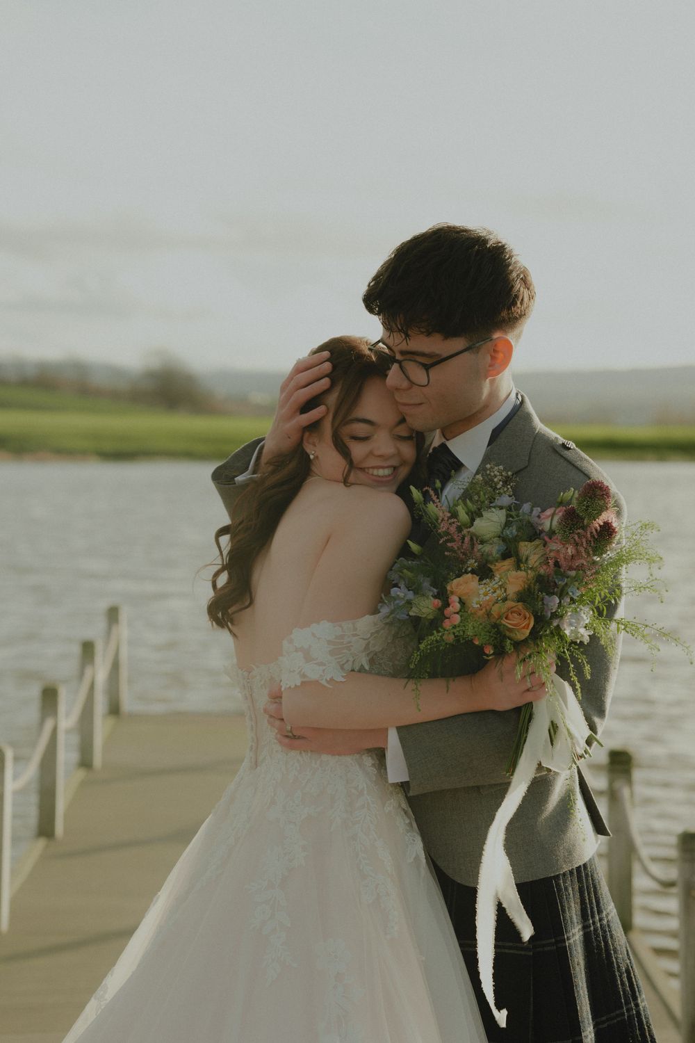 Couple embracing on wooden dock by lake, groom in kilt holding bride with floral bouquet outdoors.
