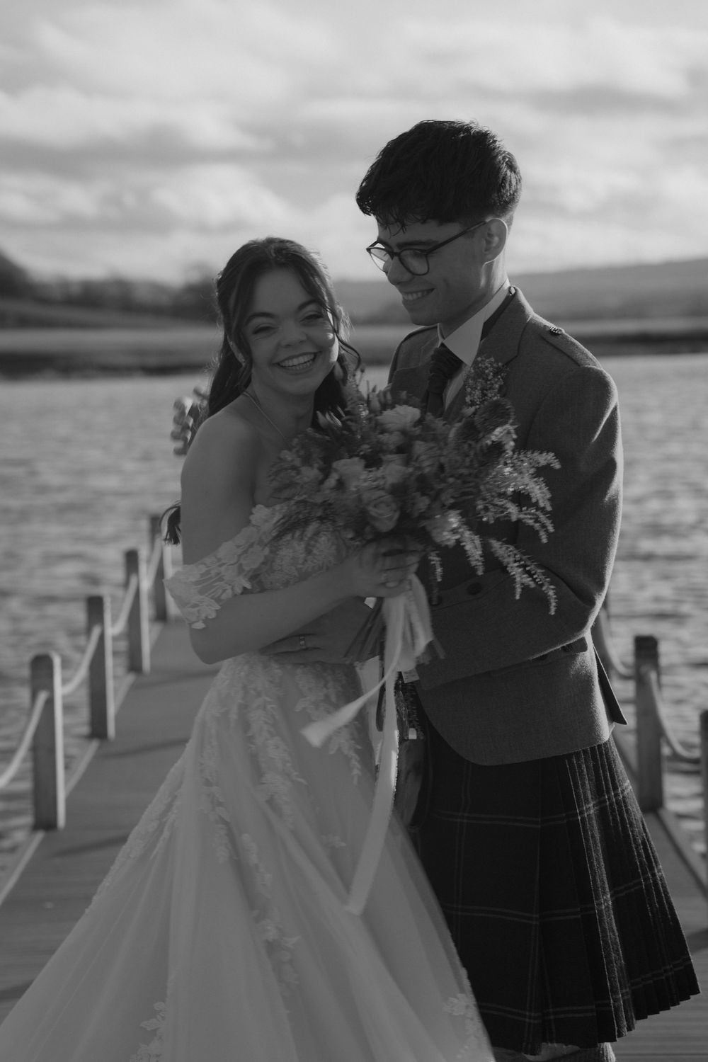 Black and white photo of a smiling bride and groom in a kilt on a wooden dock by a lake.