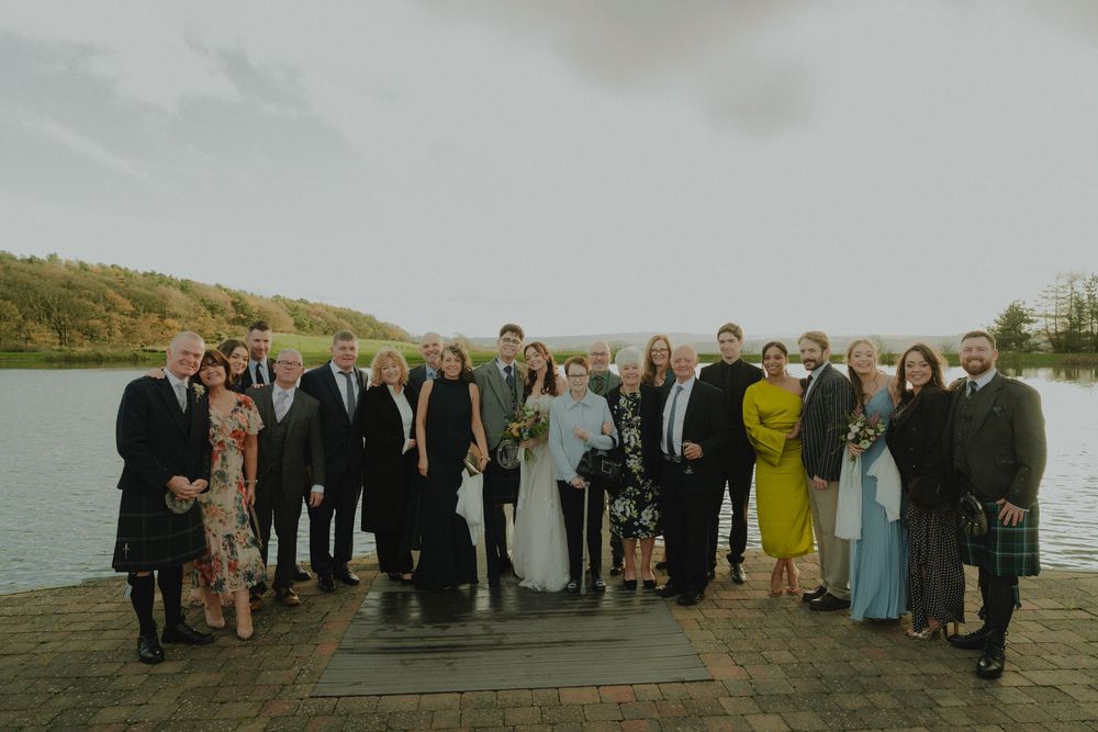 Wedding group photo of bride, groom, and guests posing on a lakeside dock with scenic autumn hills in the background.