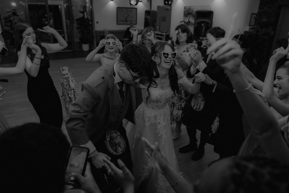 Black and white photo of bride and groom dancing joyfully with guests at a lively wedding reception.