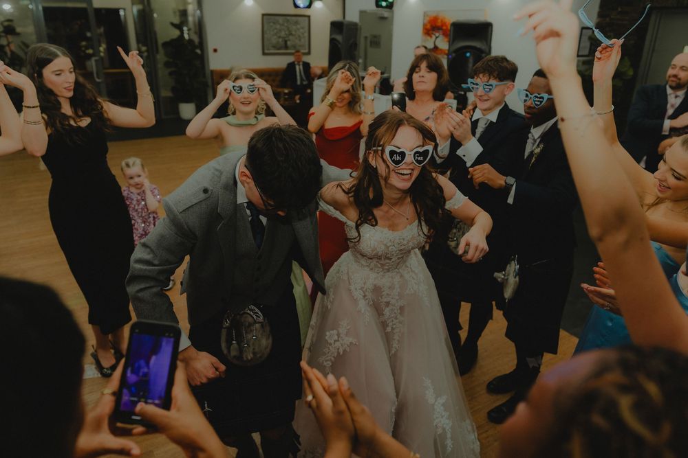 Bride and groom dancing with guests at wedding reception, wearing fun sunglasses props on dance floor.